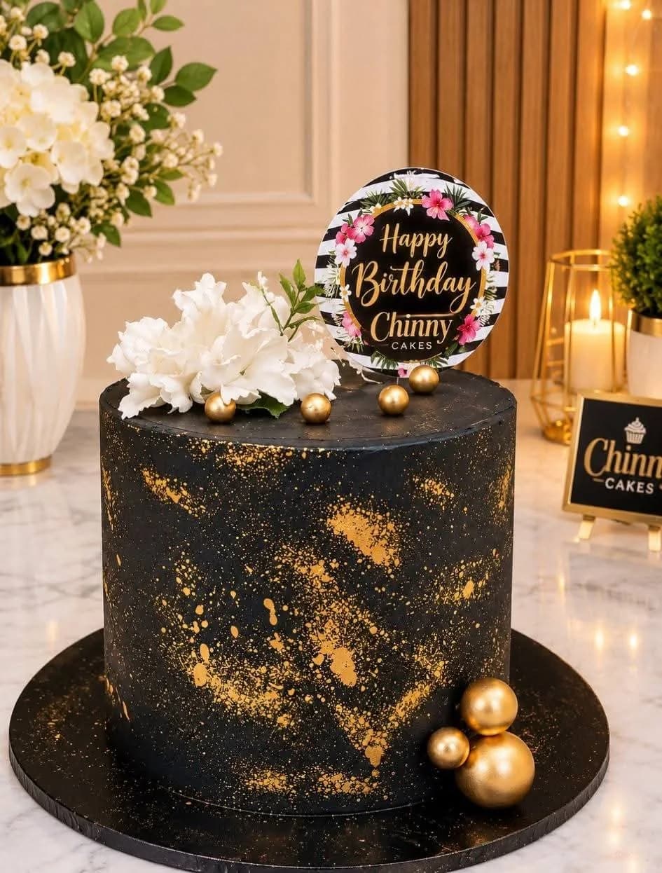 Black birthday cake with gold splatter, white flowers and gold spheres by Chinny Cakes & More